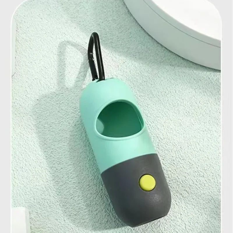 A light teal capsule dog waste bag dispenser with a black handle and a yellow button, resting on a white surface.