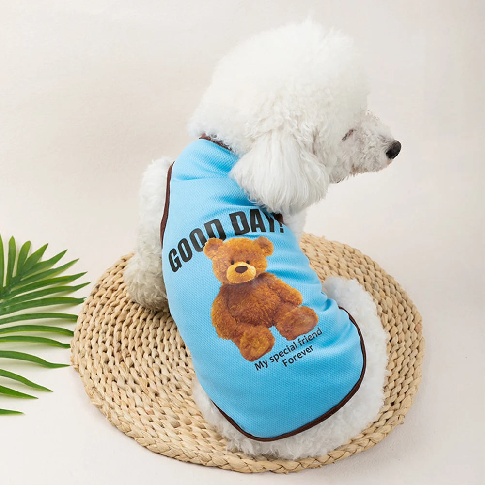 A white fluffy dog wearing a blue vest with the text Good Days and a teddy bear print, resting on a woven mat.