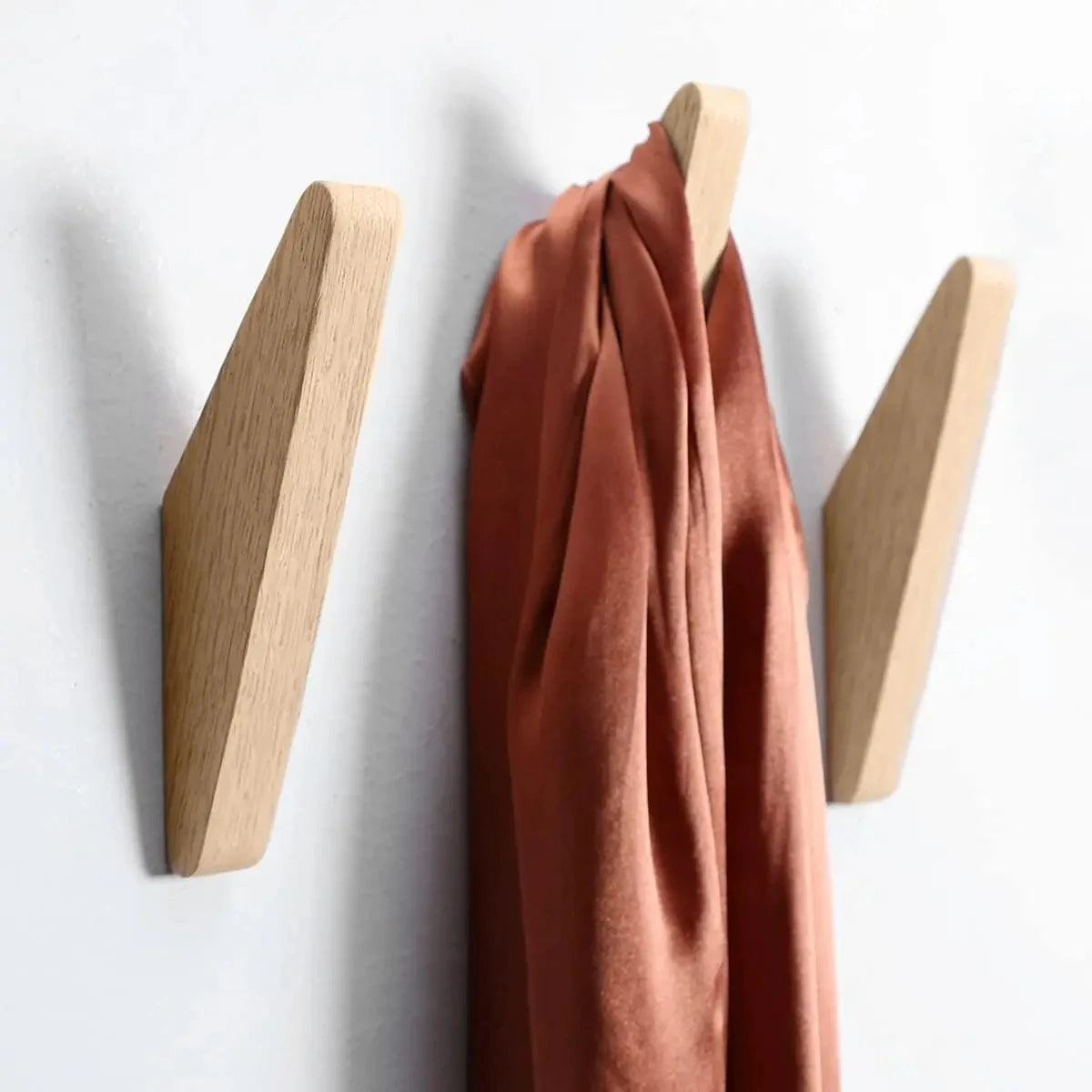 A terracotta colored fabric coat draped over an oak wood wall mounted triangle coat rack with three hooks.