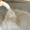 A white liquid is being poured into a gray mesh laundry bag inside a washing machine drum.