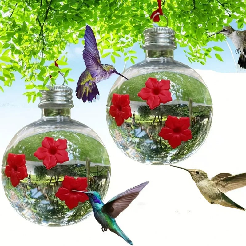 Glass hanging bird water feeder filled with water and red flowers, surrounded by green leaves and hummingbirds.