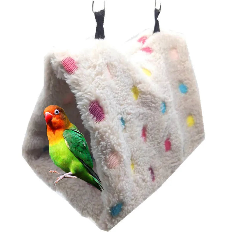 A small green and orange parrot is perched inside a fluffy, light gray plush hammock bed.