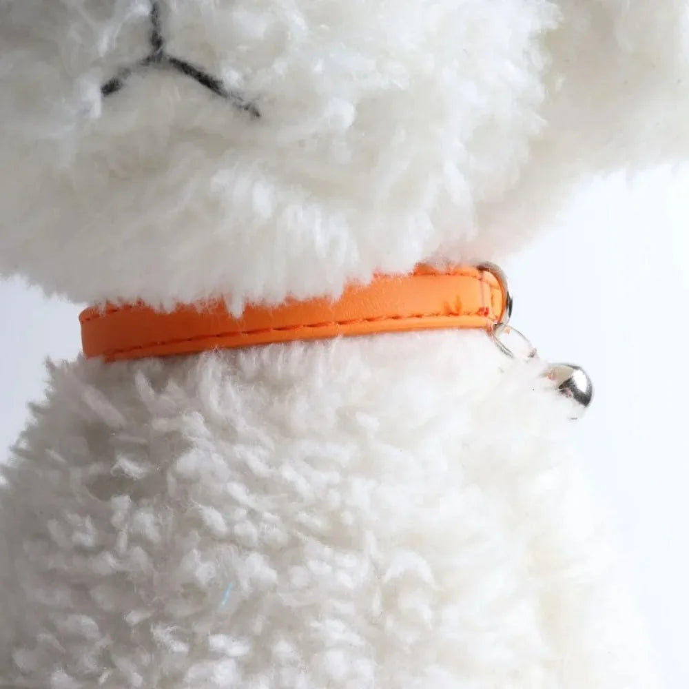 An adjustable cat collar made of soft orange PU leather with a metal bell attached, shown against a white fluffy background.