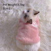 A fluffy white and gray kitten wearing a pink embroidered vest on a white, furry surface.