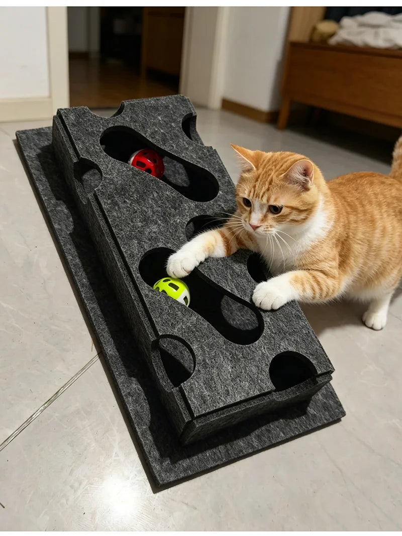 A ginger cat is interacting with an interactive puzzle toy box featuring a maze tunnel and a red ball.