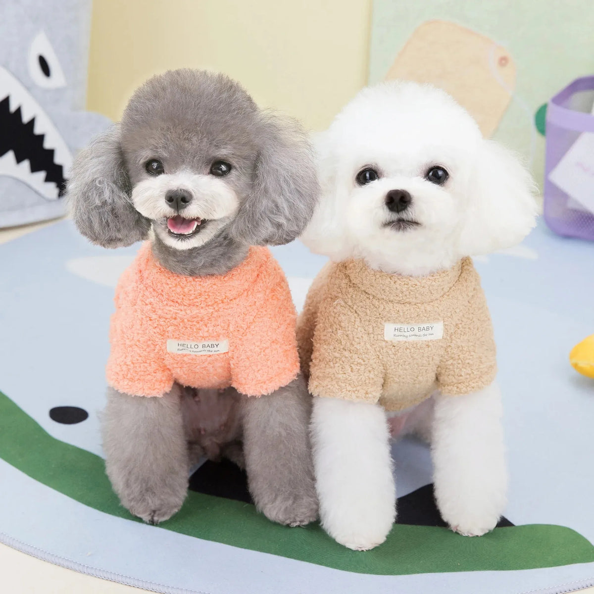 Dos pequeños perros de peluche parecidos a poodles vistiendo chaquetas suaves de forro polar, uno gris y otro blanco.