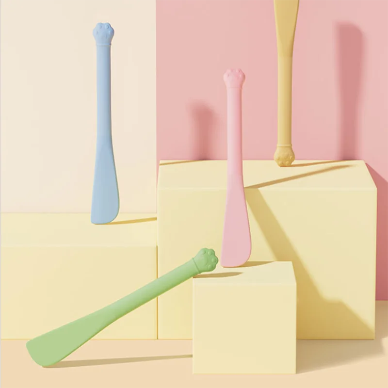 A pink pet feeding spoon is displayed on a yellow surface next to a light blue one, set against a pale pink background.