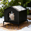 A black insulated outdoor cat house with an arched window displaying a cat inside, set on a wooden surface covered in snow.