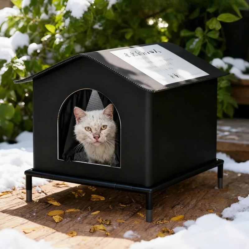 A black insulated outdoor cat house with an arched window displaying a cat inside, set on a wooden surface covered in snow.