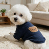 A white poodle wearing a navy blue waffle knit dog shirt with a tan bear towel embroidery, sitting on a cream rug.