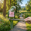 A green parrot standing on a branch next to a person's hand on a dirt path surrounded by greenery.
