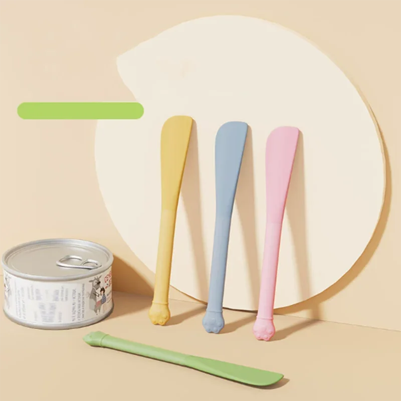 A set of five multi-purpose pet feeding spoons in various pastel colors, including a green one, displayed against a light bac