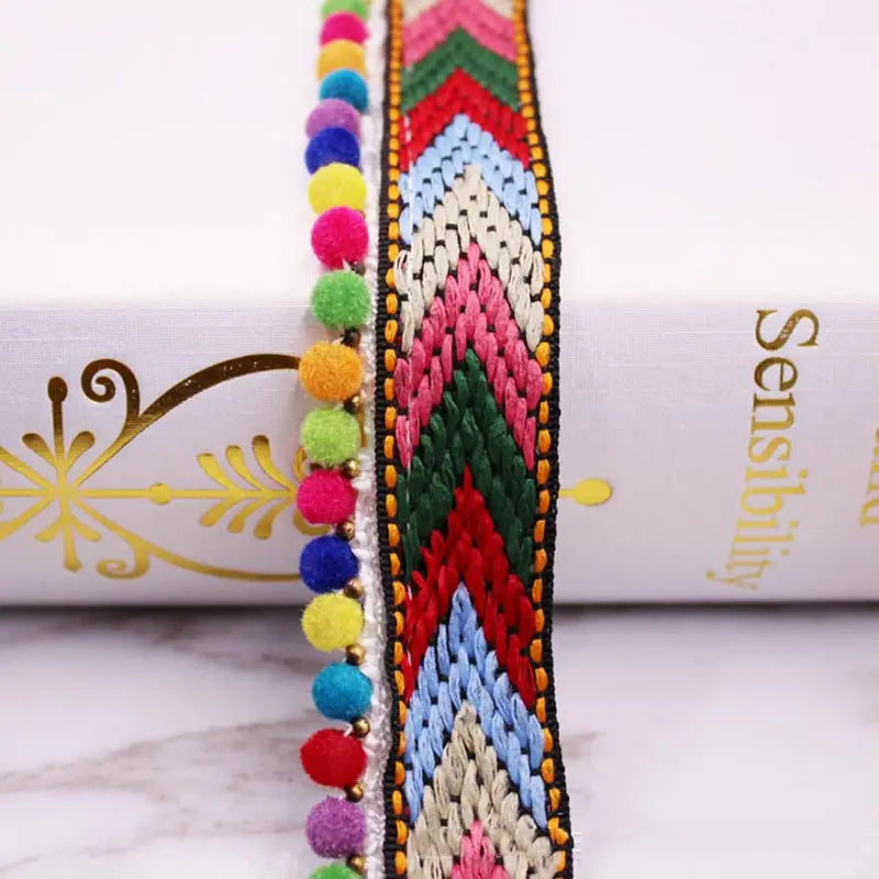 Close-up of a colorful mini pearl pom pom fringe lace trim with geometric patterns on a white fabric background.