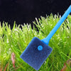 A blue dual-sided aquarium cleaning brush is shown resting on bright green aquatic plants.