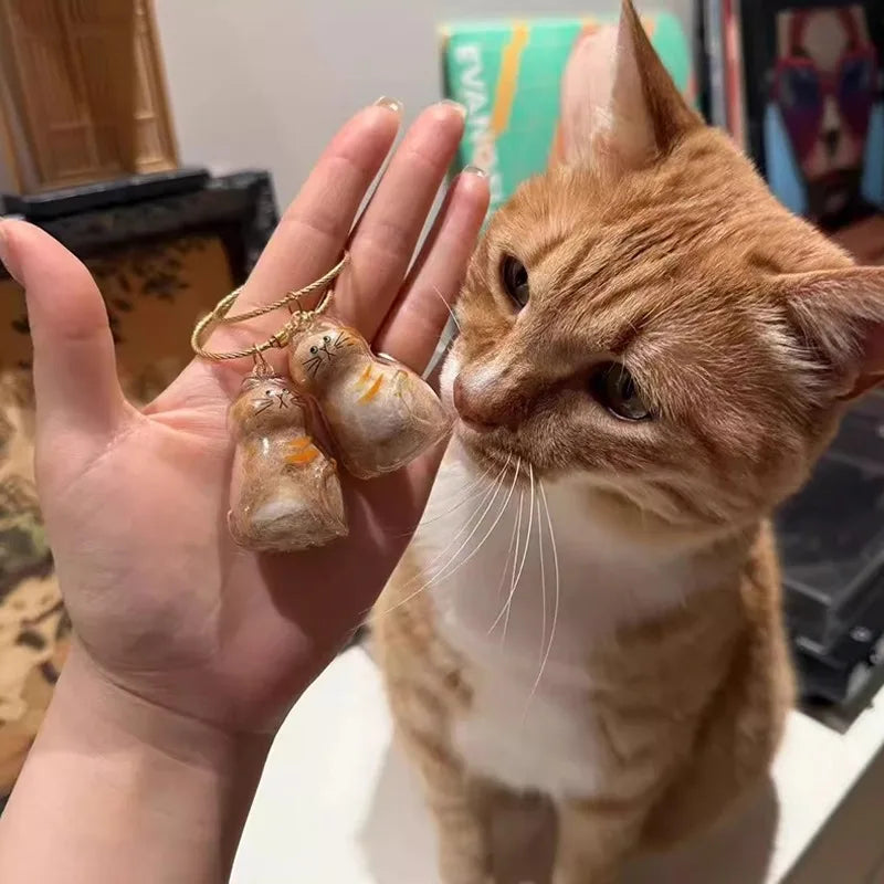 A ginger cat is looking at a hand holding two small, amber-colored fur charms on a gold chain.