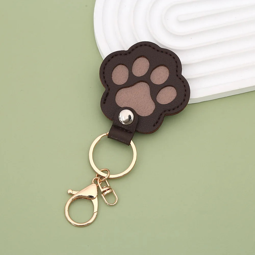 A dark brown leather paw print airtag keepsake attached to a gold-colored keyring, set against a light green background.