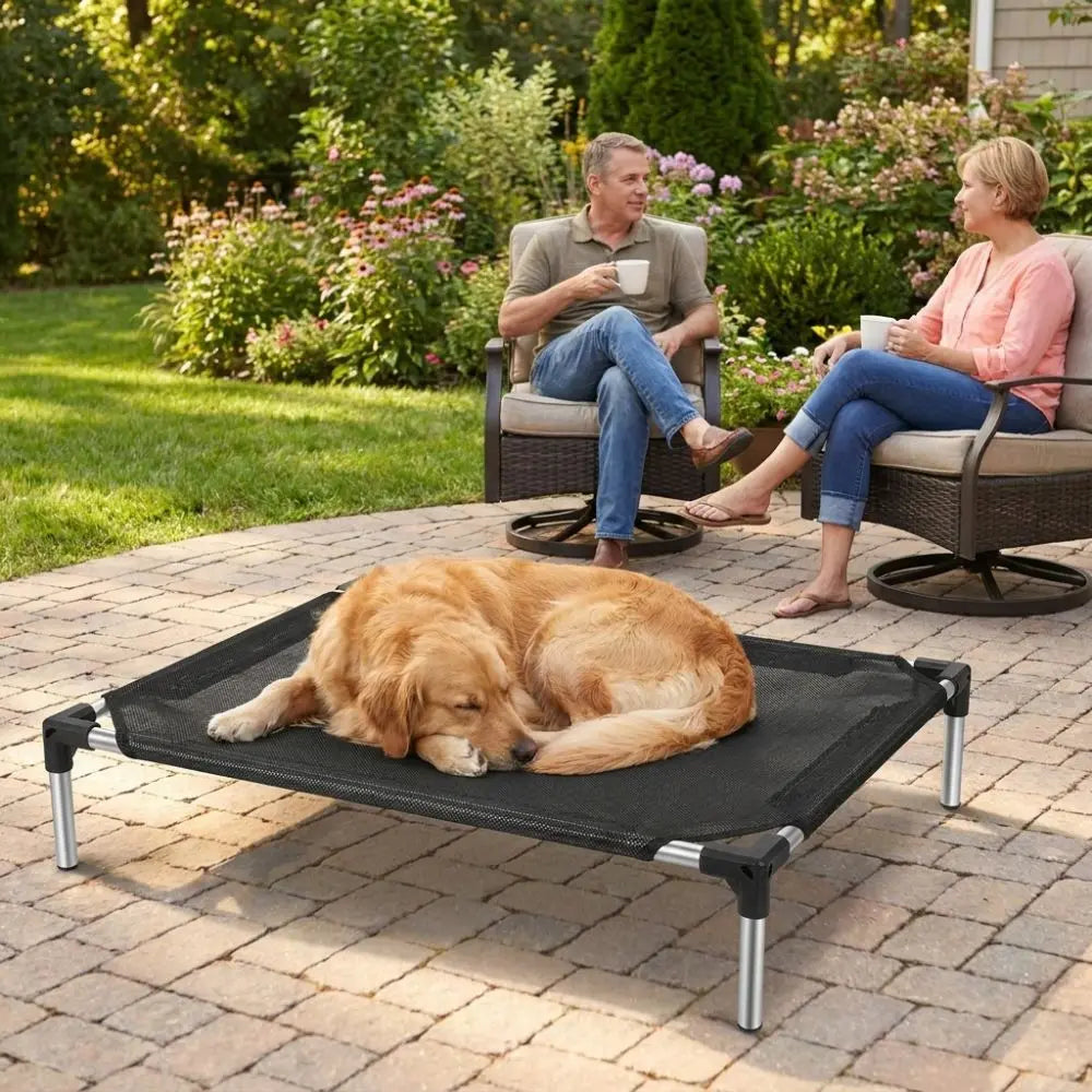 A golden retriever rests on an elevated mesh pet bed outdoors with two people relaxing nearby.