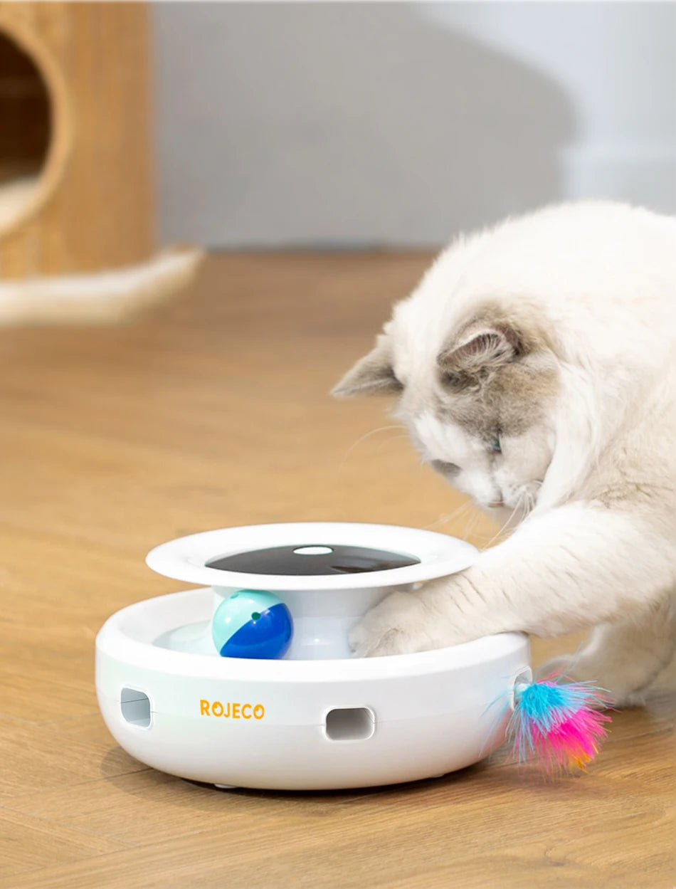 Eine weiße, automatische interaktive Spielzeug mit blauer Kugel und rosa Feder wird von einer Katze auf einem Holzfußboden ge