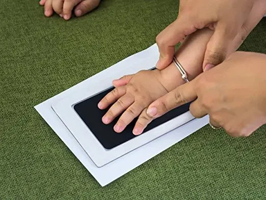 Kit d'empreinte de main montrant l'empreinte d'un bébé sur du papier blanc, avec une main humaine qui guide l'empreinte.
