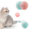 A fluffy gray cat is looking at two pink and teal electronic pet balls, with smaller versions shown nearby.