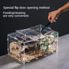 A hand is using a tool to open the flap door of a transparent acrylic reptile terrarium for feeding or cleaning.