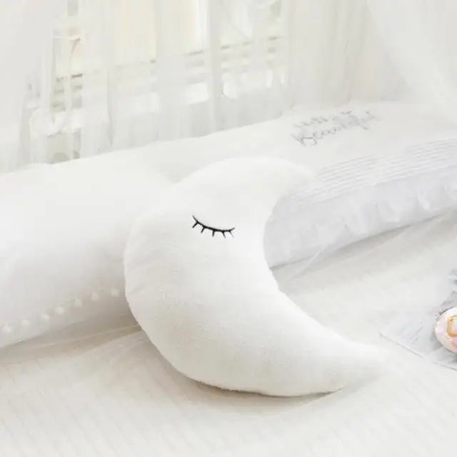 A white crescent moon shaped plush pillow resting on a white bed with sheer curtains.