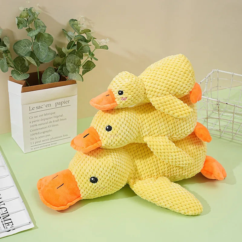 A yellow plush duck toy with orange feet and a squeaker is displayed on a green surface next to potted greenery.