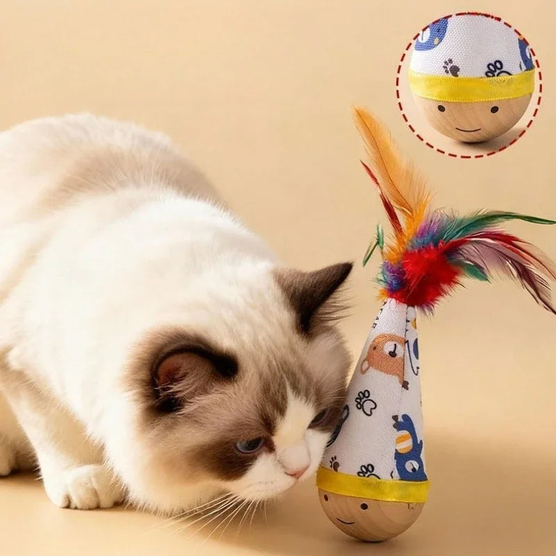 A fluffy white and gray cat is looking at an interactive toy featuring a colorful feather and a small wooden figure.