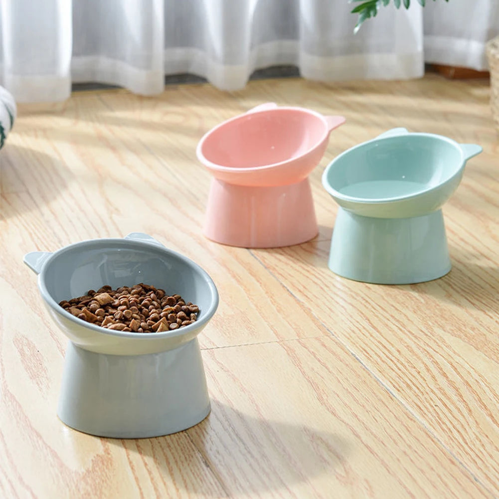 Three tilted ceramic cat food bowls in pink, mint green, and gray sitting on a light wooden floor.