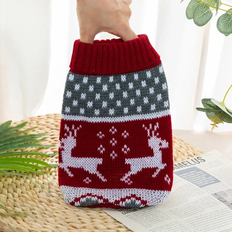 A knitted dog sweater in red and gray patterns is held by a hand, displayed on a woven mat.