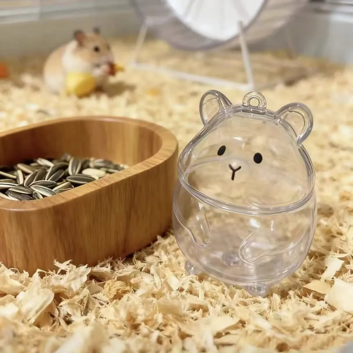 A clear plastic hamster figurine is placed on wood shavings next to a wooden food bowl and a small rodent.