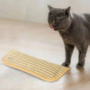 Eine graue Katze schaut auf ein beiges rechteckiges interaktives Kratzspielzeug auf einem Holzfußboden.
