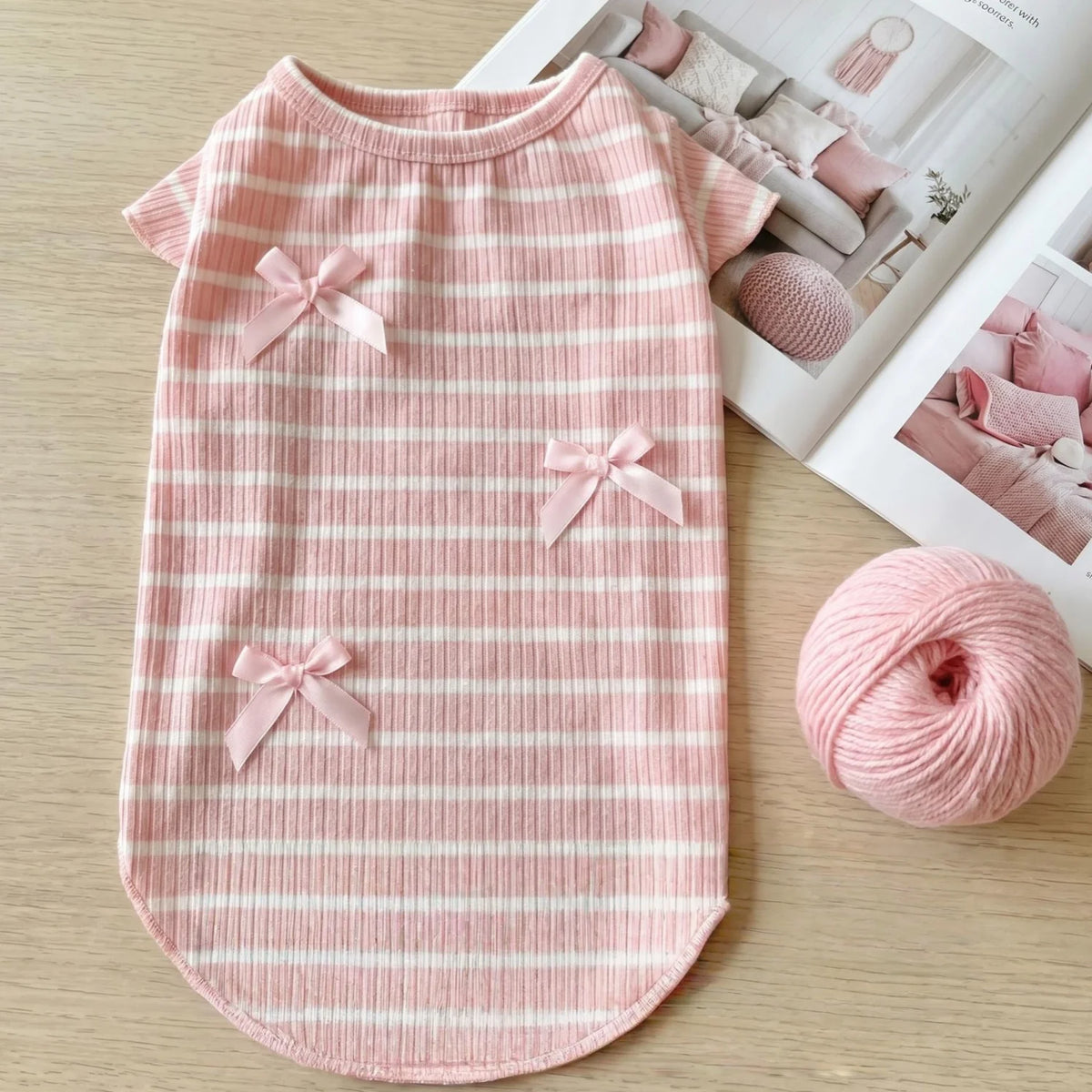 A pink and white striped knit pet shirt with decorative bows lying on a light wooden surface.