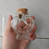 A clear glass heart-shaped jar with a cork stopper and a silver pendant featuring a feather is held in a hand.