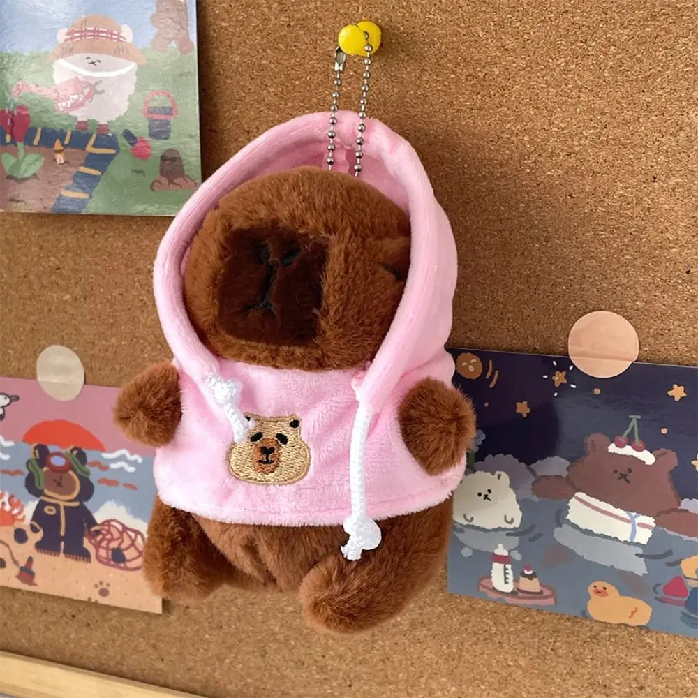 Braune Capybara-Plüschpuppe mit pinkem Hoodie und weißem Rand, neben Karten