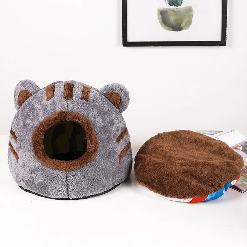 Grau-brauer flauschiger Bärenkopf Katzenbett mit Eingang neben einem braunen, runden Stoffstück.
