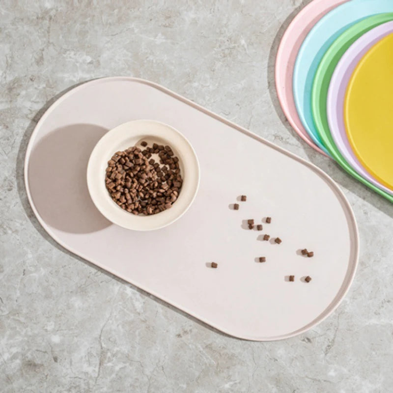A light pink non-slip feeding mat with a small bowl of dry pet food and a stack of colorful plates nearby.