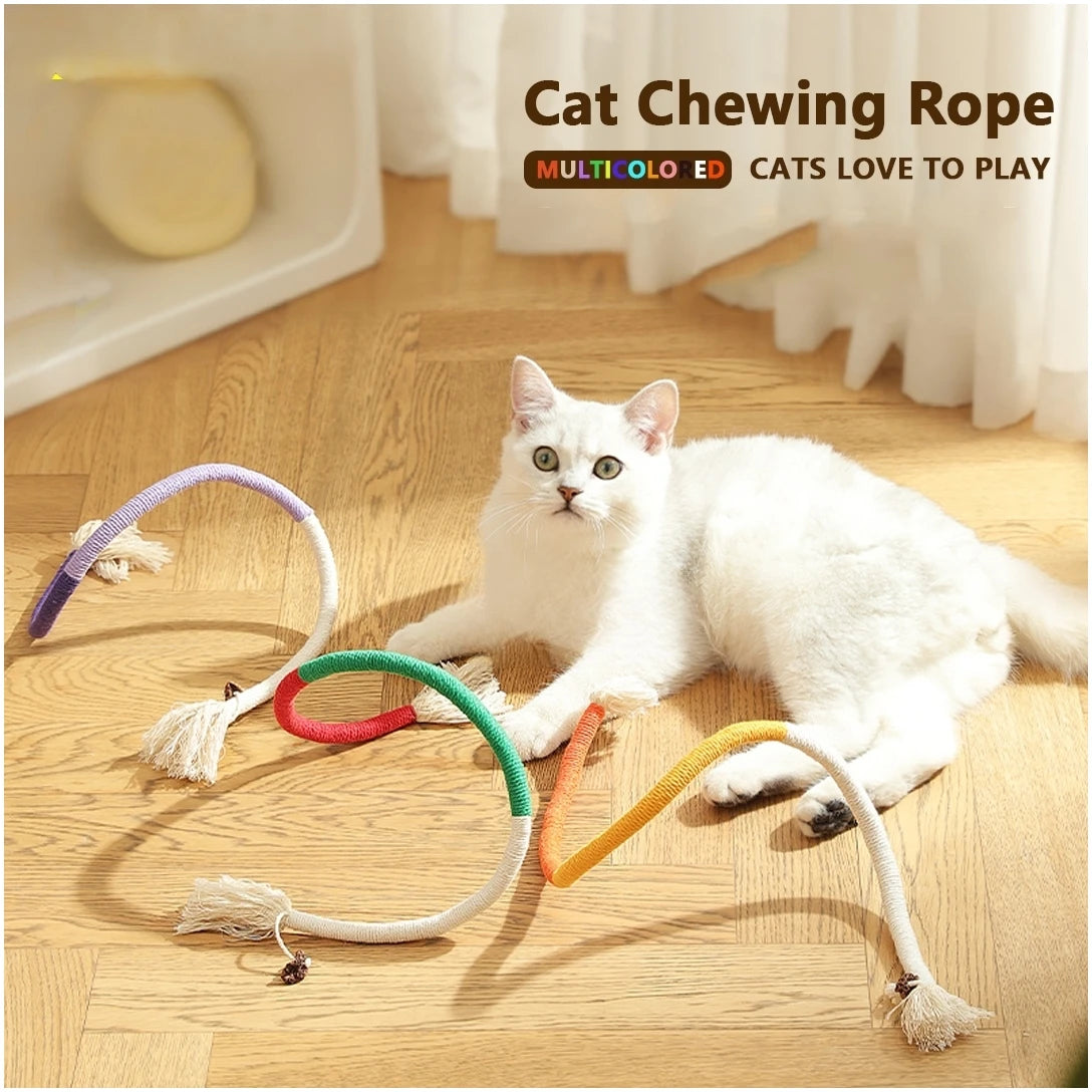 A white cat playing with various colored cat chewing rope toys on a wooden floor.