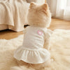 A fluffy light beige dog wearing a white knitted dress with pink heart details, sitting on a cream rug.