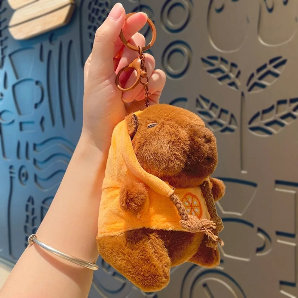 Eine Hand hält eine braune Capybara-Plüschpuppe mit orangefarbener Kapuzenjacke und goldfarbener Halskette.