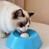 Un gato blanco interactuando con un cuenco de alimentación lenta azul, lamiendo una pelota blanca interna.