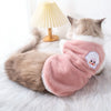 A fluffy gray cat wearing a pink embroidered vest on a white fur rug next to a lamp.