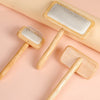 A wooden pet grooming brush with massage bristles is shown lying on a light pink surface.