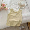 A pale yellow knitted mini vest with lace trim is displayed on a white surface next to pink roses and a white bow.