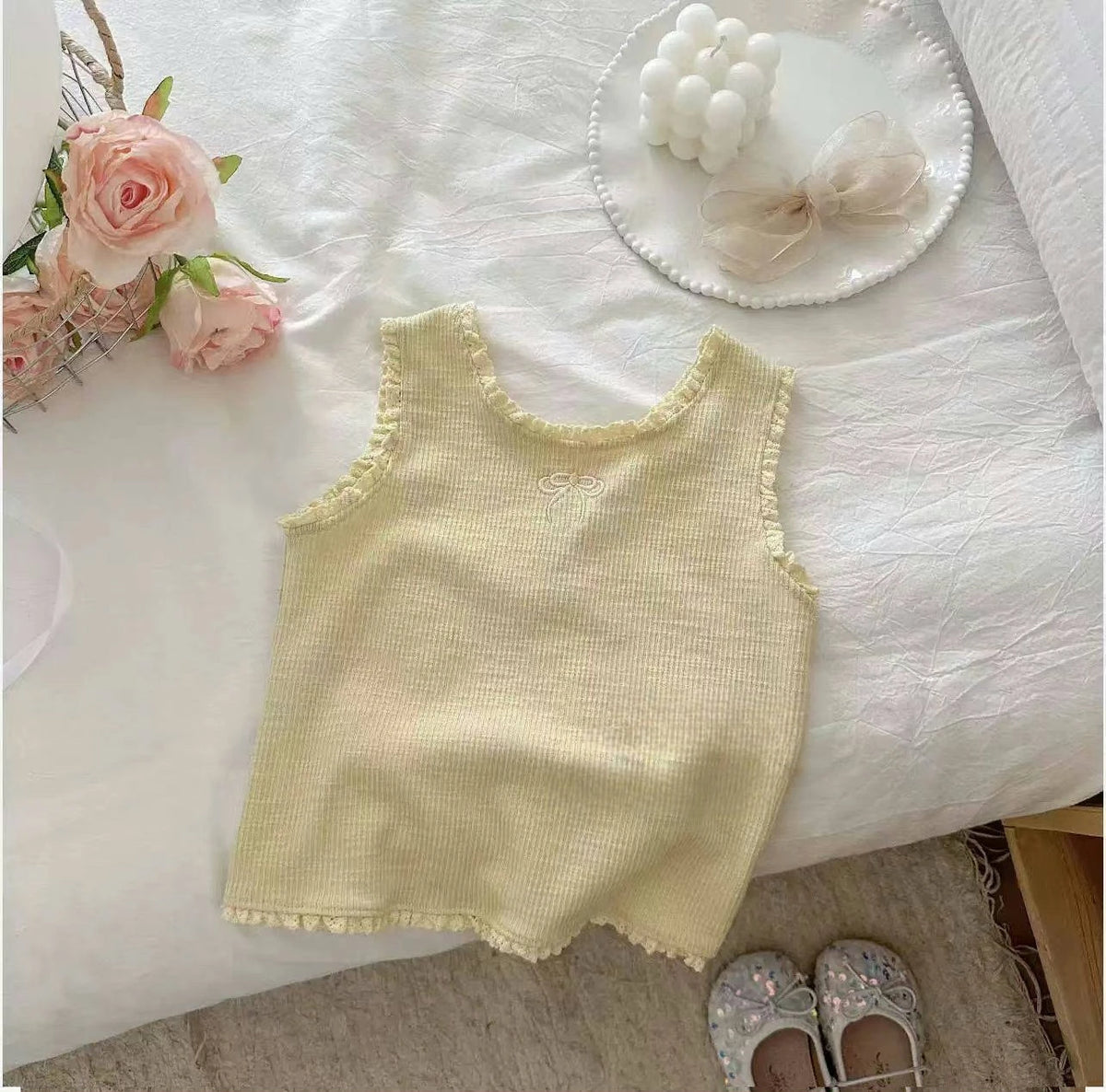 A pale yellow knitted mini vest with lace trim is displayed on a white surface next to pink roses and a white bow.