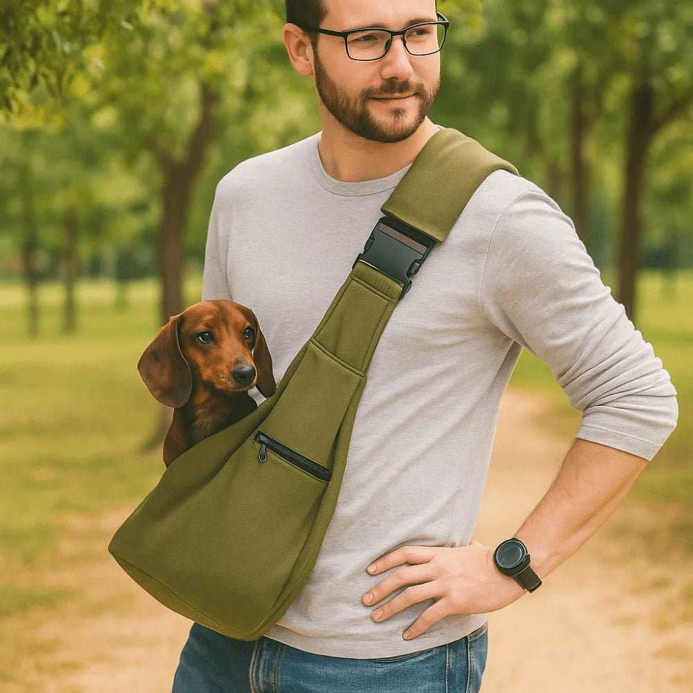 A man wearing a light gray shirt holds a olive green pet carrier bag with a safety harness and a dog in a park setting.