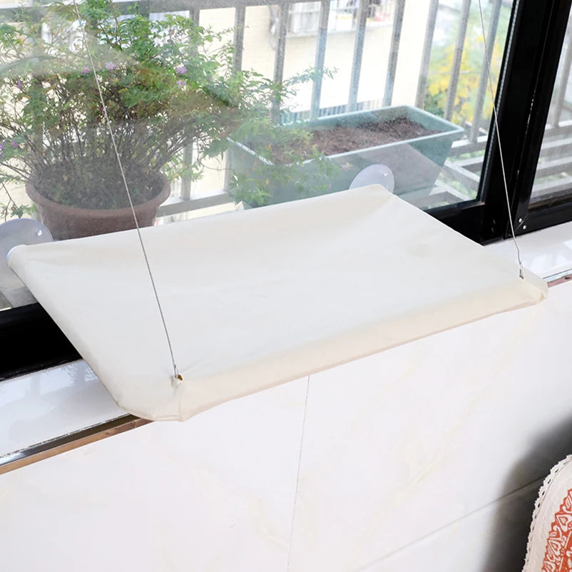 Un lit-hamac pour chat blanc fixé à une fenêtre est exposé sur un rebord de fenêtre avec de la verdure en arrière-plan.