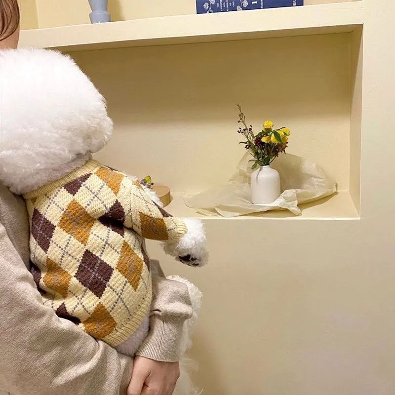 A person is holding a luxury plaid knit sweater for dogs with white faux fur trim, standing in front of a light-colored shelf