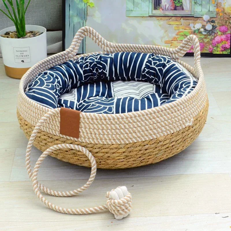 A woven rattan cat bed with a removable cushion and a rope handle is shown on a light wooden floor.