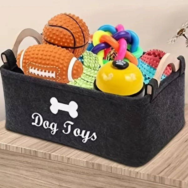 A black felt pet toy storage basket filled with various colorful dog toys including a ball, bone, and ring toys.